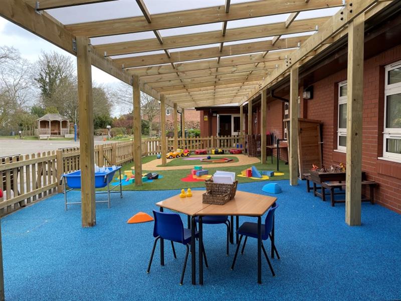A school table with three blue chairs has been placed onto blue wetpour surfacing underneath a timber canopy, with artificial grass installed behind the blue wetpour. The canopy is attached to a school building. 