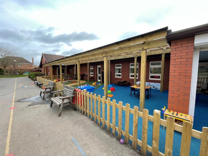 Timber canopy attached to the school building with bow timber fencing around it, blue wetpour surfacing, artificial grass and a roadway made out of wetpour all underneath the canopy. 