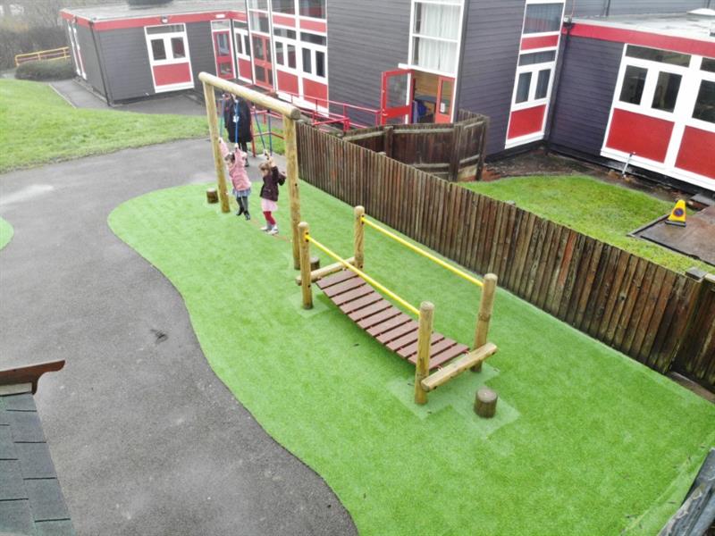 Two children moving across a three piece timber trim trail, including a clatter bridge, stepping logs and a rope traverse installed onto artificial grass surfacing