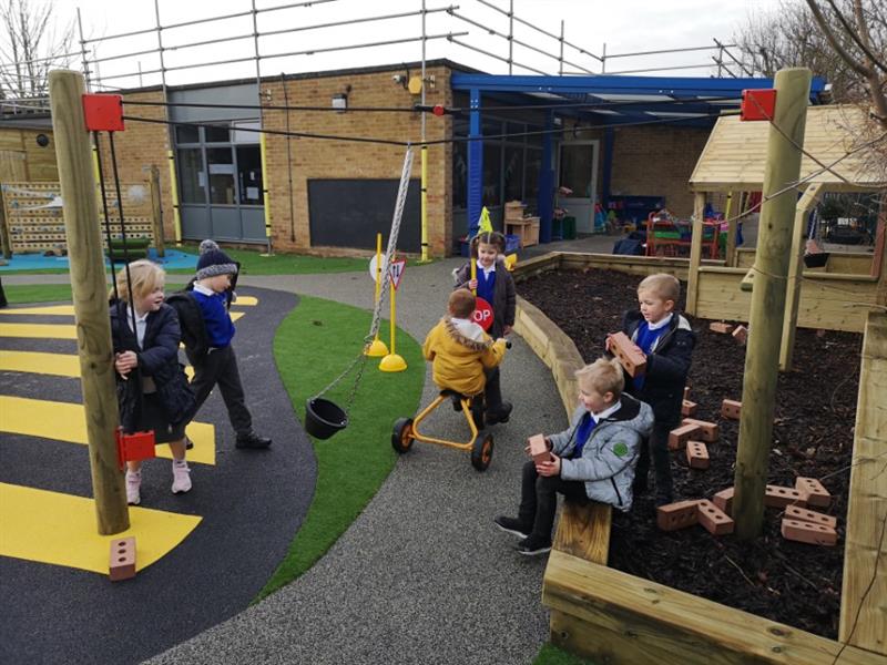 6 children playing on playground, with one child wearing a yellow coat riding a trike, two children holding bricks and two more children playing with a rope and pully. 