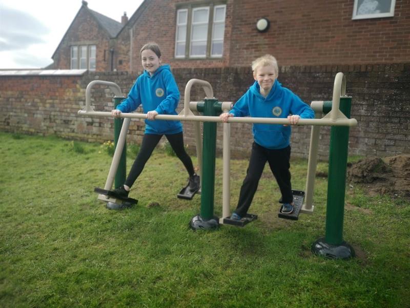 2 children, one boy with blonde hair and one girl with brown hair are playing outside on outdoor gym equipment which has been installed onto the school field next to the school building. 