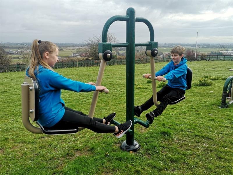 2 children wearing blue school jumpers, one boy with brown hair and one girl with brown hair, the girl has black trainers on with a pink tick, are playing on the outdoor gym equipment in the school field with a view of fields and houses behind them. 