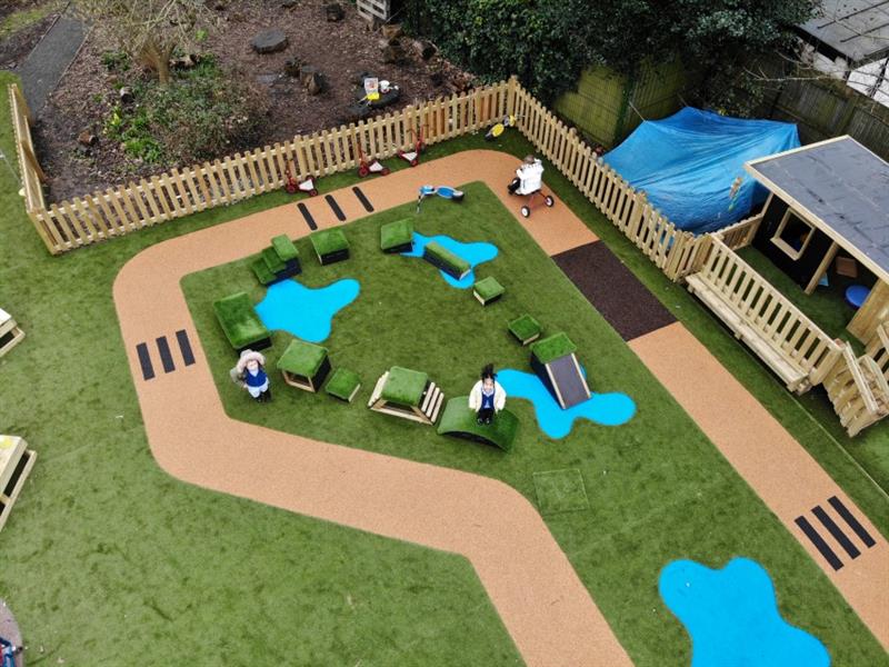 Aerial view of eyfs playground whilst one child wearing a white coat rides a trike on the roadway, one child wearing a pink coat stands on a get, set go! block! and one child stands on artificial grass next to the blocks. 