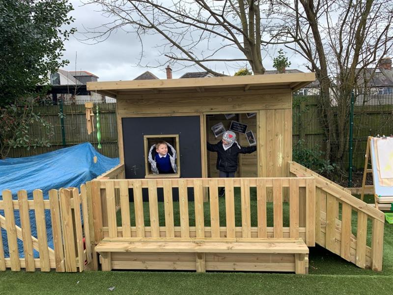 2 children, one wearing a grey coat and one wearing a black coat and grey hat are stood inside of the lookout cabin smiling, cabin has been installed in front of school fencing. 