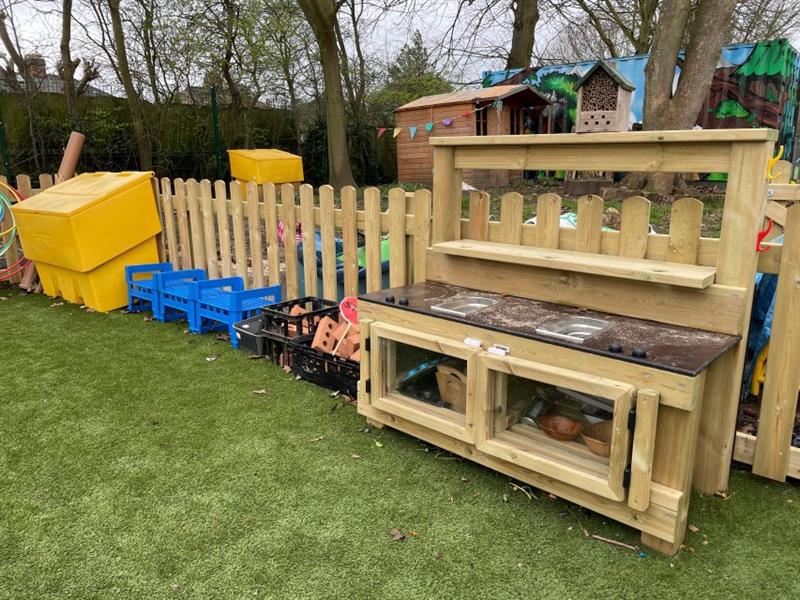 A mini mud kitchen has been installed onto artificial grass in front of bow top timber fencing, next to the kitchen are some black boxes containing bricks, blue crates and very large yellow boxes. 