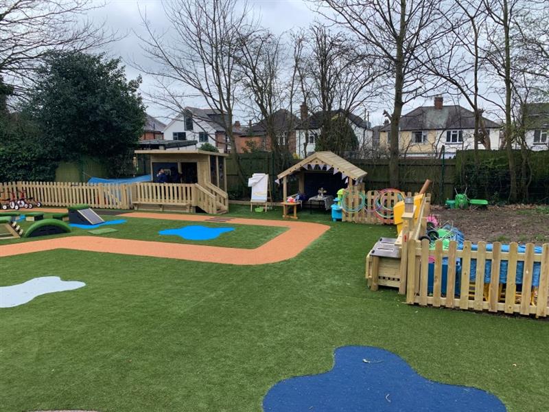 Landscape photograph of school playground which includes bow timber fencing, artificial grass, a roadway made out of wetpour surfacing, a giant playhouse and a lookout cabin. 2 children stood outside of the lookout cabin. 