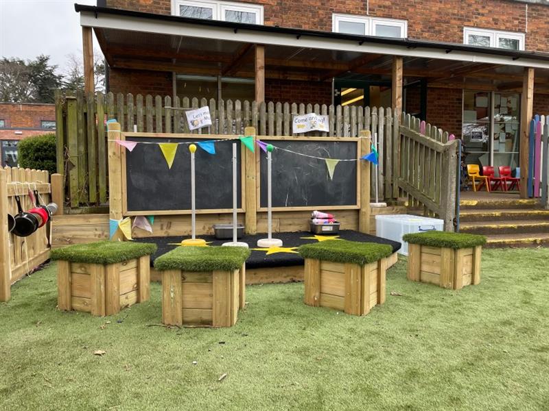 performance stage with 2 chalkboards installed onto artificial grass in front of the school building with 4 artificial grass topped seats in front of the performance stage for children to sit on. 