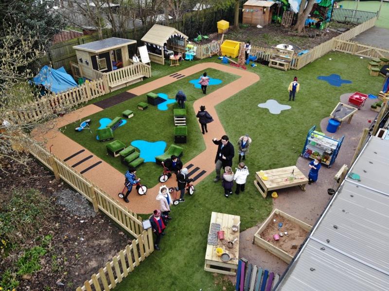 15 children playing in an eyfs playground, 3 on trikes, 1 in the lookout cabin, 1 at the construction table whilst one teacher wearing a black coat supervises. 