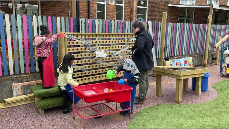 2 children playing with a red water box and green watering can whilst one child stands on artificial grass topped steps pouring water into the water wall whilst one teacher wearing a black coat watches. 