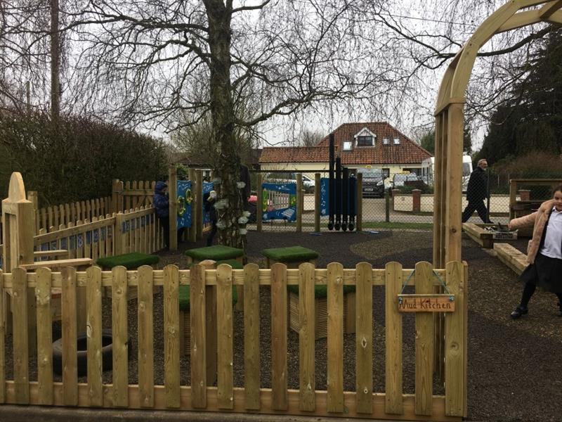 The new timber fence in front of the storytelling area and music area in the new outdoor space, with a small sign attached to the fence saying mud kitchen. Inside the area is a girl wearing a pink coat and a boy wearing a blue coat. 