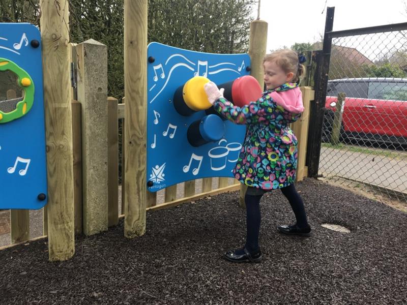 One girl wearing a navy coat with pink and green flowers on, banging on red, yellow and blue bongo pannel posts that have been installed onto earth brown coloured play bond surfacing.