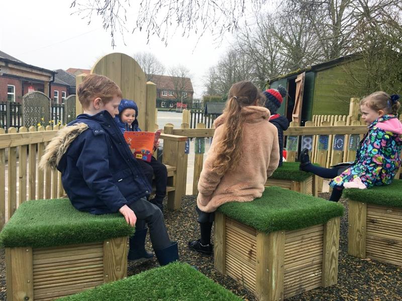 4 children sat on moveable artificial grass topped seats in front of a boy sat on a storytelling chair with the school building in the background.