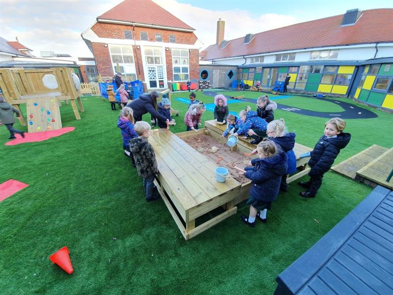 12 children playing in the sand box with one teacher supervising, sandbox has been installed onto artificial grass near the school building, with one child smiling at the camera. 