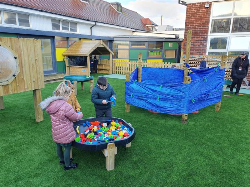 3 children playing with different toys on a table that has been installed next to the modular play tower and den making posts onto artificial grass with one teacher supervising. 