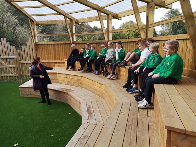 Close up shot of 13 children sat on the seats in the amphitheatre listening to instructions from one teacher wearing a purple coat. 