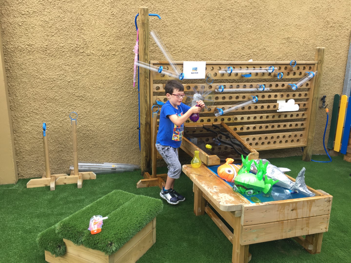 A little boy is playing with a Water Wall and other water play equipment on an artificial grass surfacing.