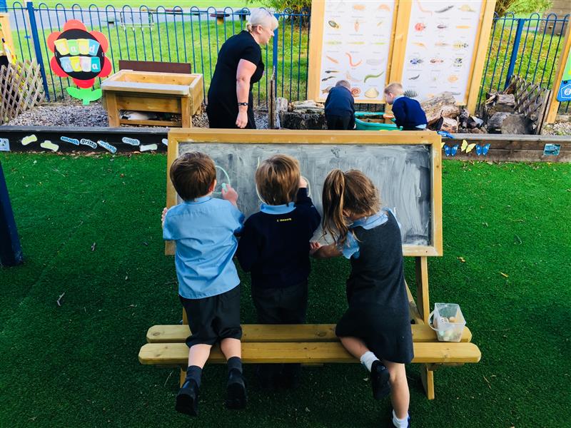 three children kneel on the double sided easel and draw on the chalkboard