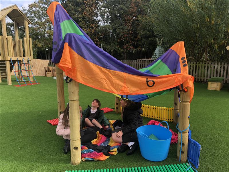 3 children sat inside a creative den they have made using 5 timber poles and an orange, purple and green sheet as a roof. They also have a large blue bucket inside the den and multicoloured sheets to sit on. 