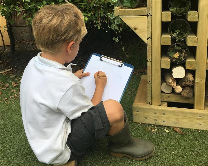 a child sits on the floor in front of the bug hotel and writes down notes on his clipboard