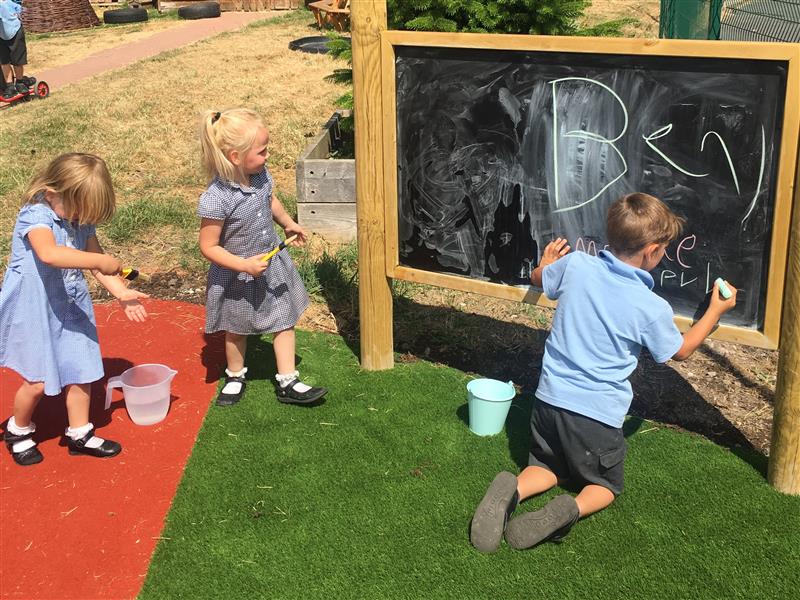 a little boy in blue and black school uniform kneels in front of the chalkboard and draws on it whilst two little girls in blue school uniform stand behind him passing him chalk