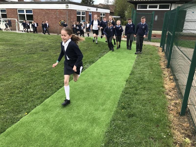 Children wearing navy school uniform are running on a daily mile track which has been created out of artificial grass and installed onto the school field which is in front of the school building. 