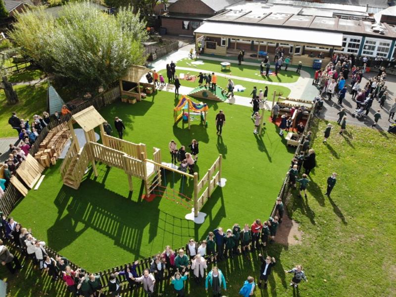 children stand around on the green artificial grass playturf that has a timber climbing frame on it and there is people gathered all around