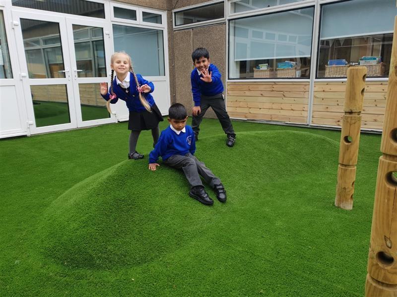 3 children wearing blue school jumpers, 2 stood on top of the playground surfacing mound whilst one child sits down on the mound which has been installed in front of the school building. 