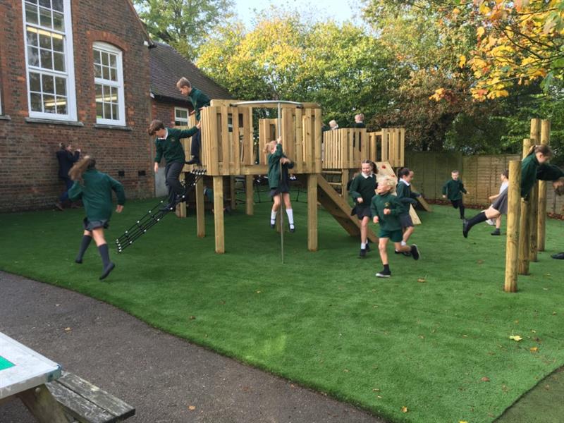 12 children wearing green jumpers are playing on the new play tower with a pole that has been installed onto artificial grass in front of the school building. One girl is sliding down the pole onto the grass. 