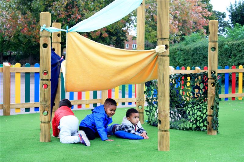 3 children lying on artificial grass under a yellow and blue sheet which they have used to make a den using the den making posts. The den has been built in front of multi-coloured fencing. 