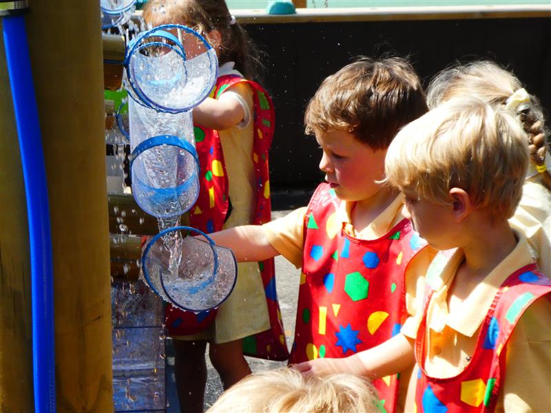 4 children, 2 boys and 2 girls wearing yellow school uniform and red aprons with multi-coloured shapes on playing with the water wall.  