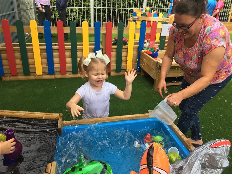 A child splashing in a water table as a teacher uses a jug to pour water down the mini damming station. The water table is full of loose play resources