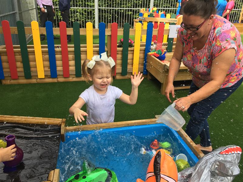 A little girl is splashing water in a water tray as a teacher is pouring water in the tub with a measuring jug. Both of them are laughing as they play