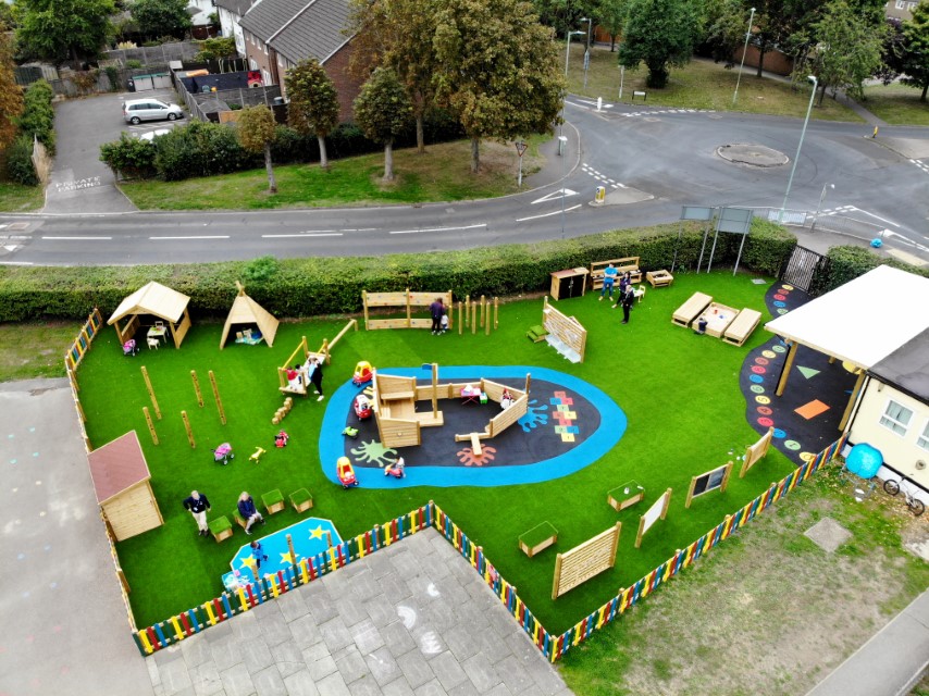 A birds-eye view of a playground with an artificial grass surface, with a multi-coloured wooden fence that goes around the play area. There is some Saferturf graphics laid on top, with a variety of play equipment around the area.