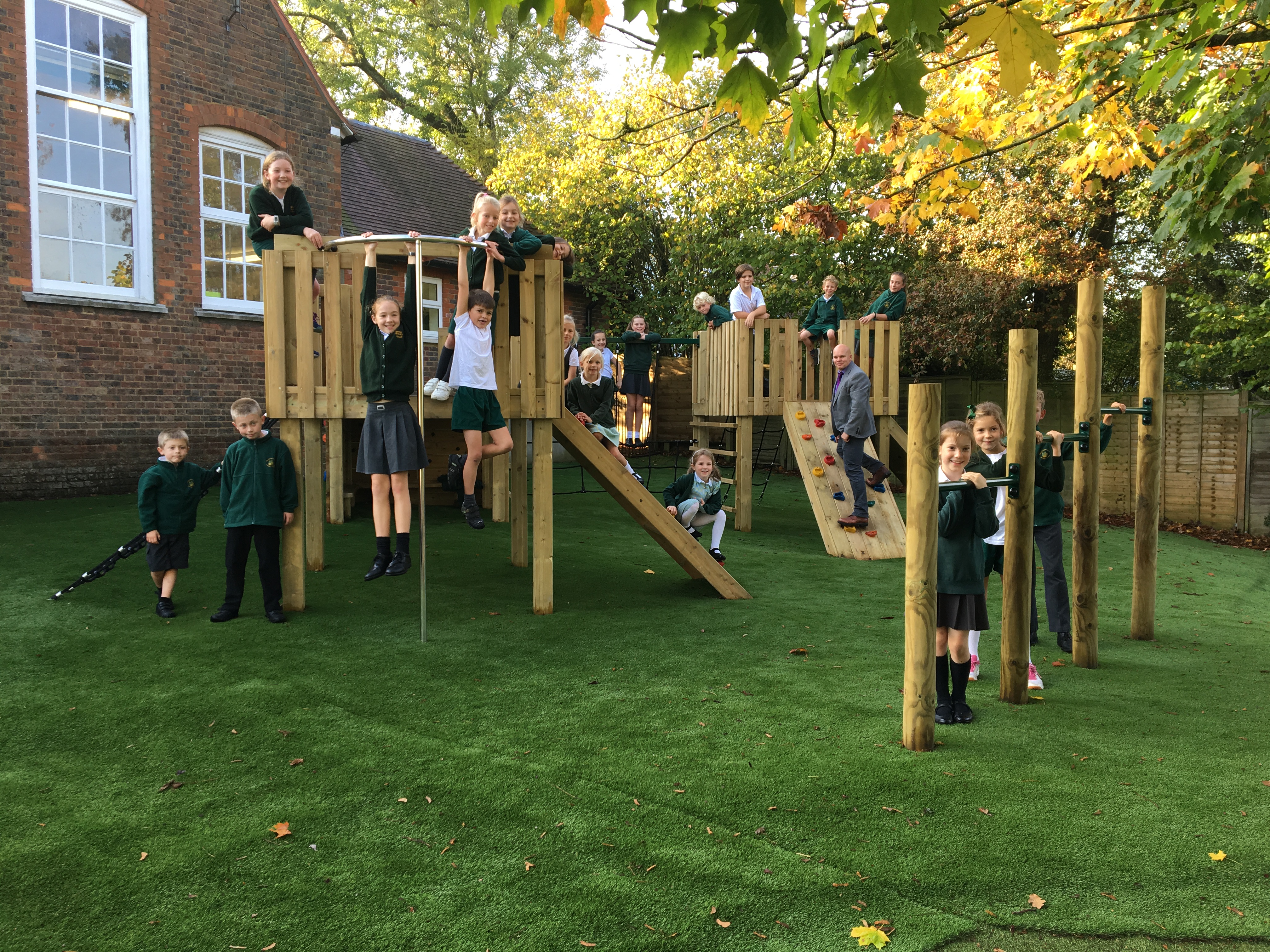 A group of children are all posing on different pieces of play equipment as they look into the camera, smiling. A teacher can be seen joining in, posing on one of the play towers with the kids. All the play equipment is wooden and installed on artificial grass.