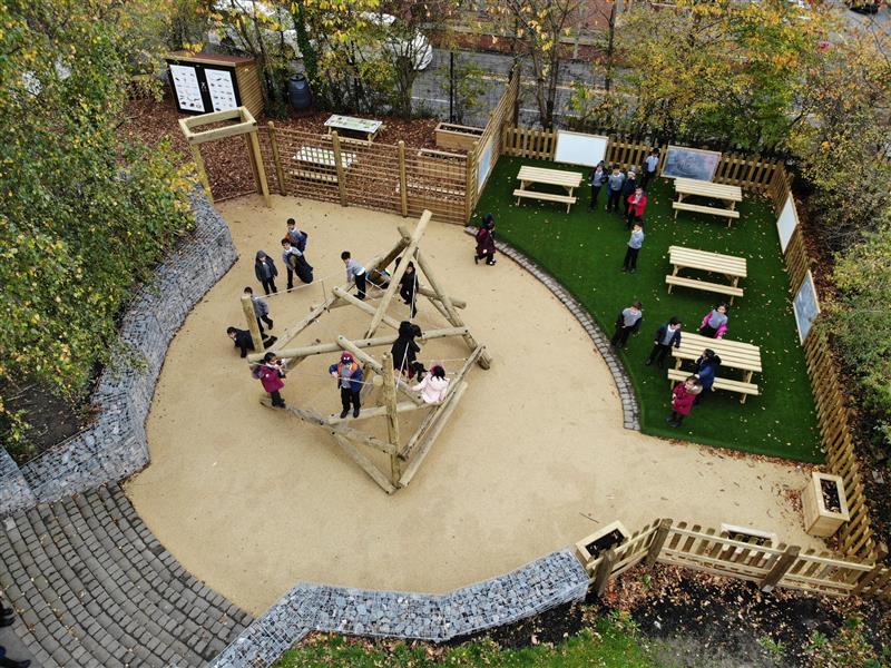 beige wetpour underneath a climber frame and artificial grass surfacing, timber picnic tables are lined  around the side with giant chalkboards and whiteboards on the fences