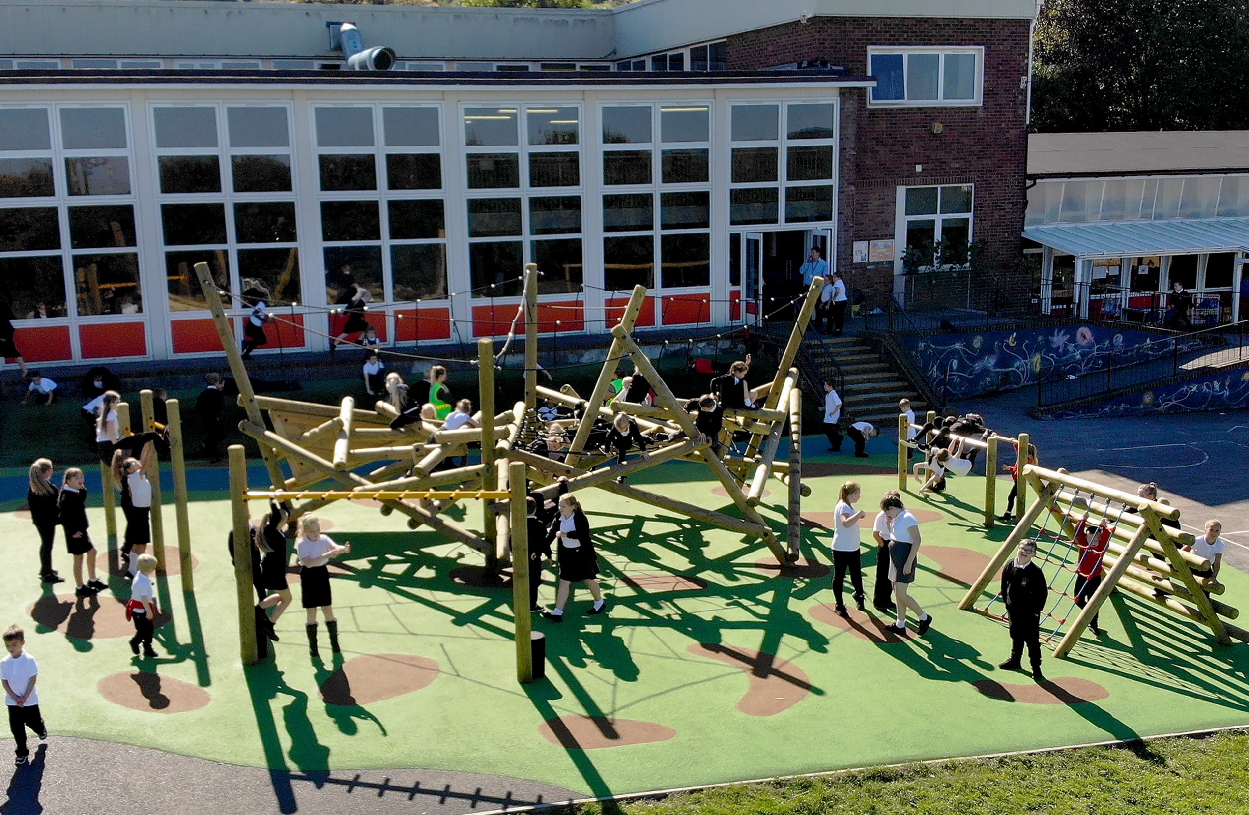 A group of children are playing on a wetpour play area, which has a huge wooden climbing frame. The climbing frame is placed facing the school building, with large groups of children running around.