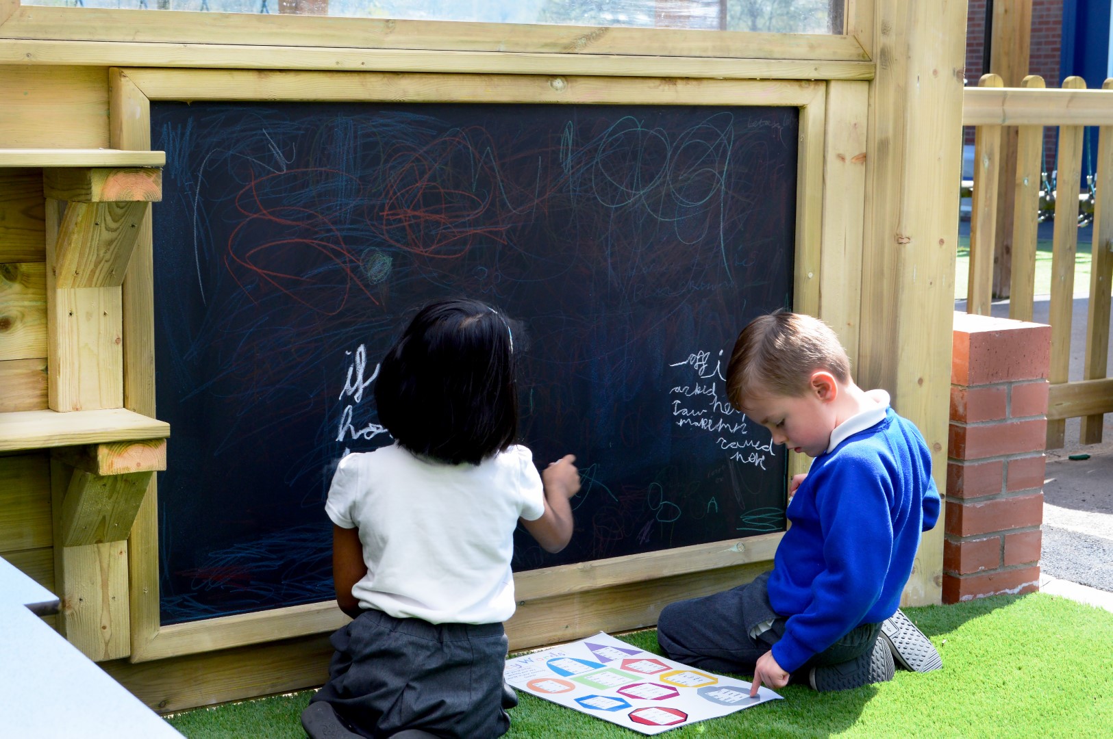 Two children sat down next to a giant chalkboard outside. Both the children are writing on the board as the boy (on the right) looks at a worksheet, laid out on the floor, for ideas of what to write and using it as a reference