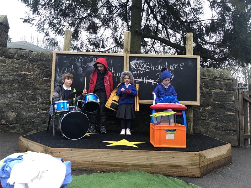 4 children stood on a performance stage in front of 2 chalkboards with scribbles on, one wearing a red coat, one wearing a blue coat and one wearing a yellow coat. 