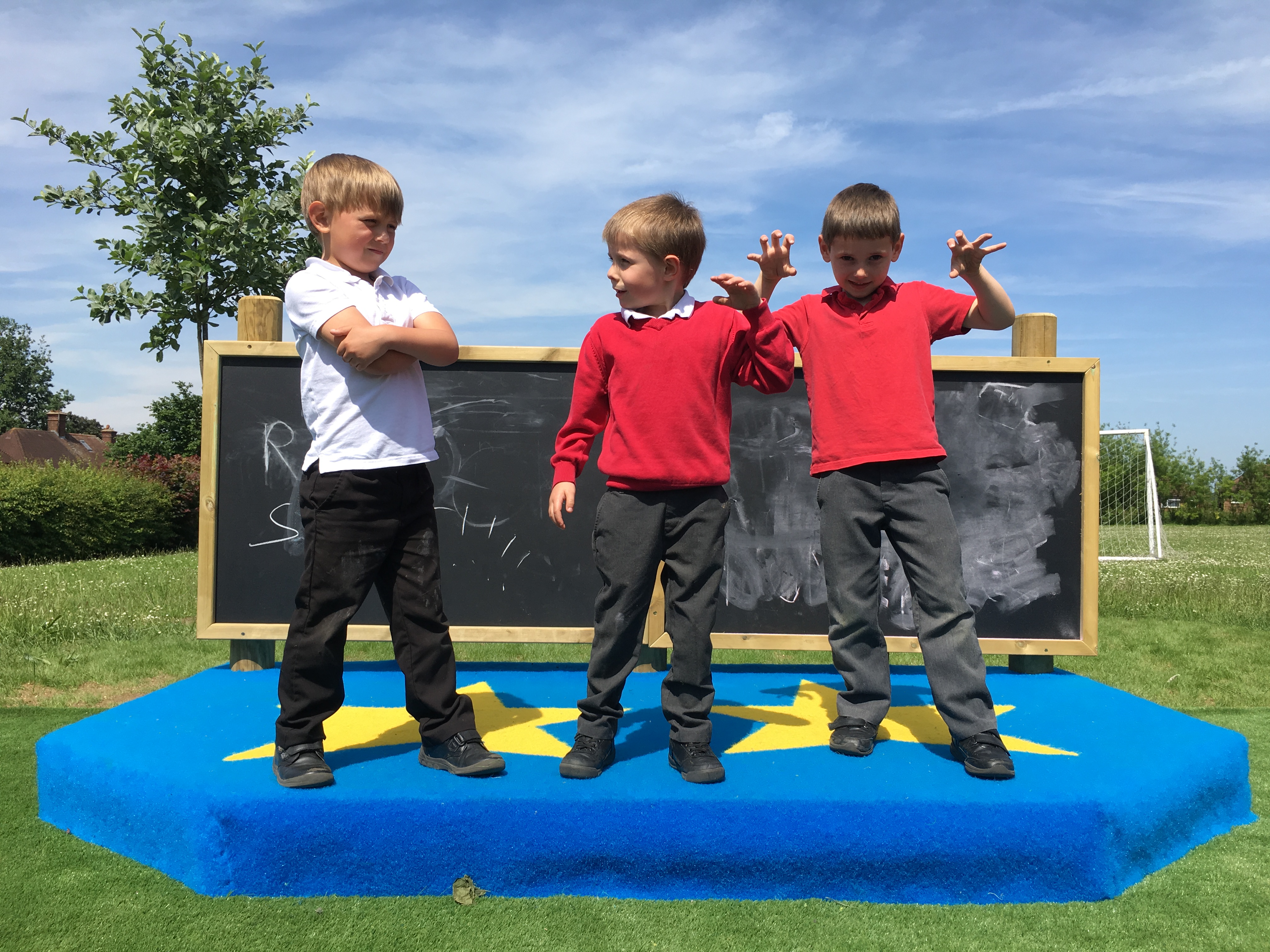 3 children are stood on a small stage, looking at the camera. 2 of the children have their arms up like their scarring something, as the other child is crossing their arms and looking disappointed in the others. Behind them is a chalkboard with marks on it. The stage is covered in blue saferturf, with yellow stars.