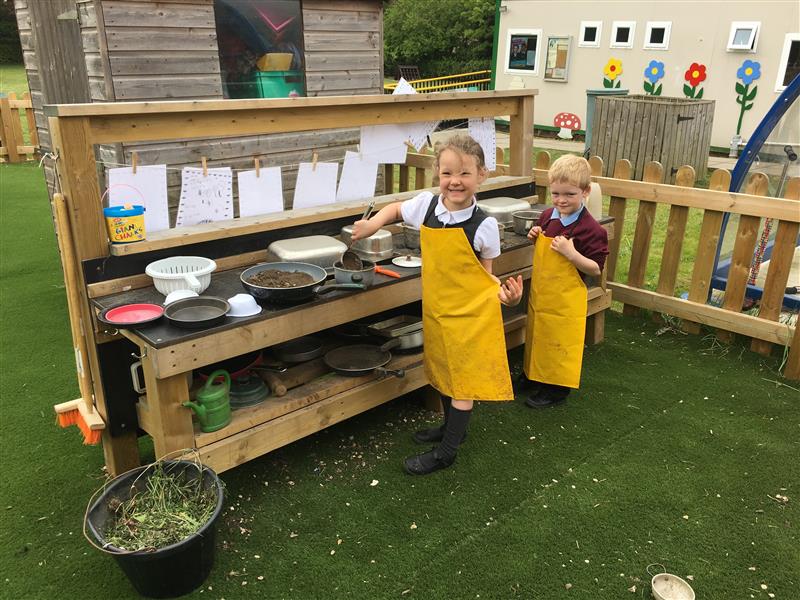 two children pose in yellow aprons as they mix mud and play in the play kitchen