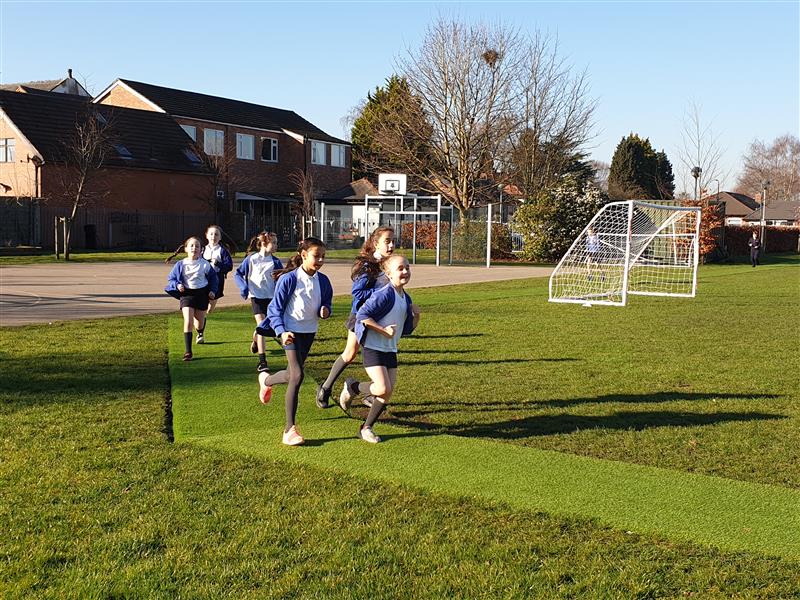 6 children, all girls wearing white polo tops and blue cardigans are running around the daily mile track which has been made using artificial grass and goes around the school field. 