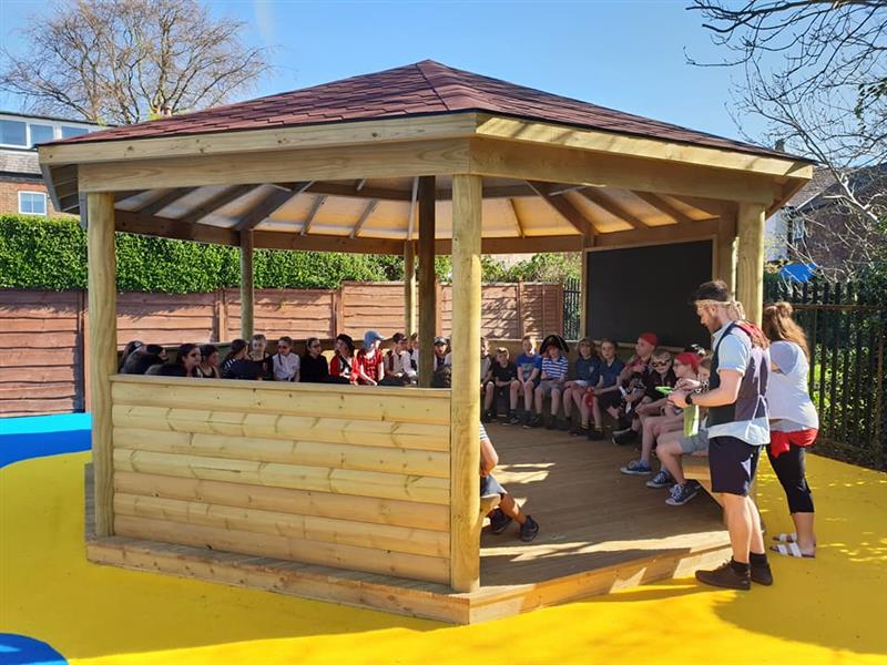 A big class of children sat inside a large hexagonal gazebo which has been installed onto blue and yellow wetpour. Children are dressed as pirates listening to 2 teachers who are also dressed as pirates. 