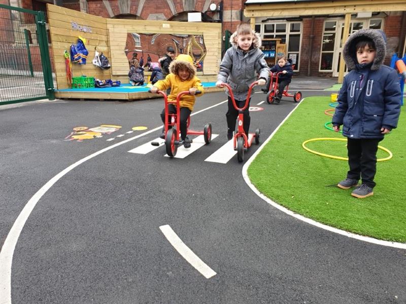 3 children riding on trikes on a roadway whilst another child wearing a blue coat stands on the artificial grass looking at the camera. 