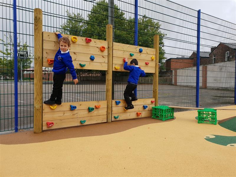 2 boys wearing blue school jumpers playing on a climbing wall that has been installed onto wetpour surfacing in front of blue fencing, with green crates placed on the floor next to the climbing wall. 