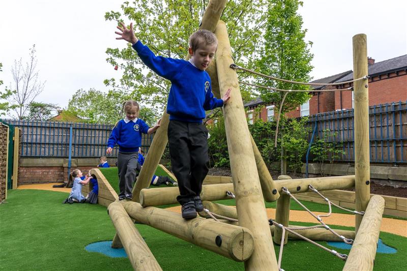 2 children, one boy and one girl wearing blue school jumpers balancing on a climbing frame that has been installed onto artificial grass whilst 5 children play in a climb through tunnel behind the climbing frame. 