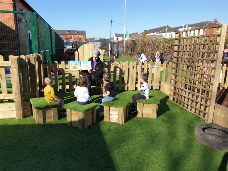children sit on grass topped seats in a semi circle facing a teacher in a story telling chair reading them a story book