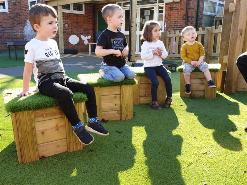 4 nursery children sat on artificial grass topped seats listening to a story being read