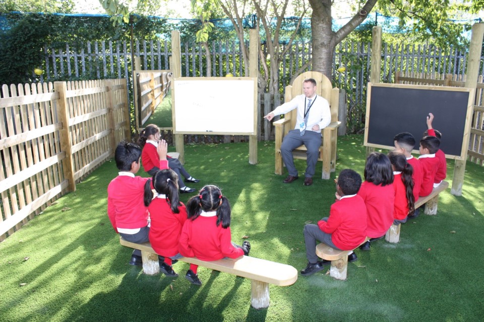 A teacher is sat on a freestanding storytelling chair as he points to a child. The children are sat on wooden benches opposite to the teacher. All the seating is outdoors, with a whiteboard and a chalkboard beside the freestanding storytelling chair.