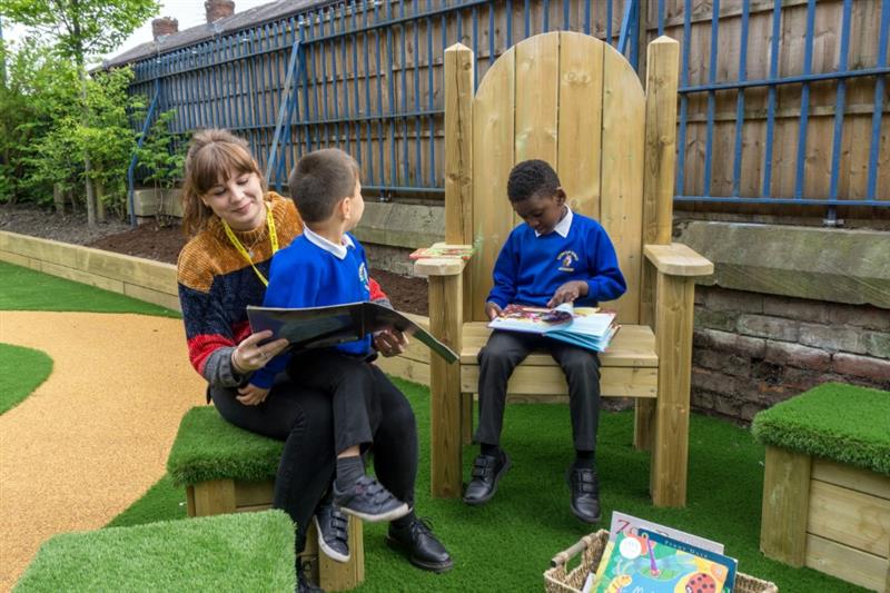 one child wearing a blue school jumper sat on a storytelling chair reading a book whilst another child sits on a teachers knee reading book. 