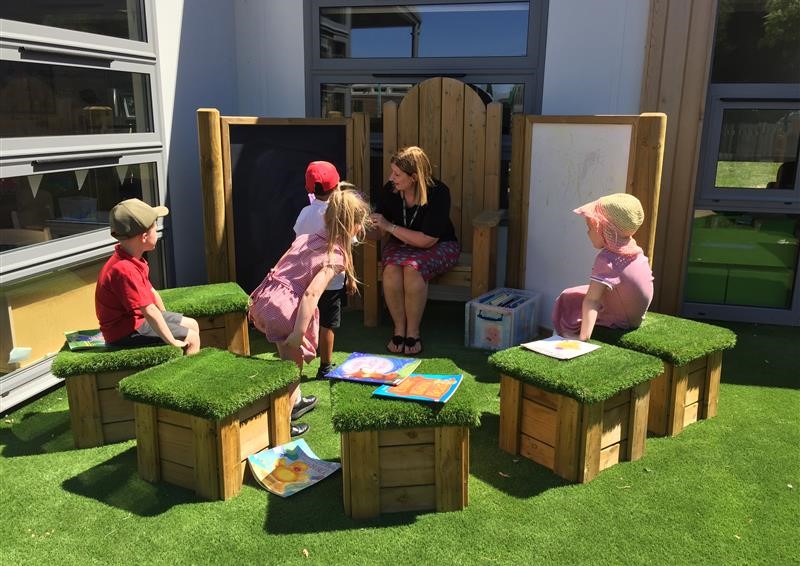 A teacher is sat on a big wooden chair, which is placed in front of 6 wooden cubes with artificial grass placed on top of them. 4 children are taking part in a lesson with the teacher, as a child approaches the chalkboard which is next to the chair.